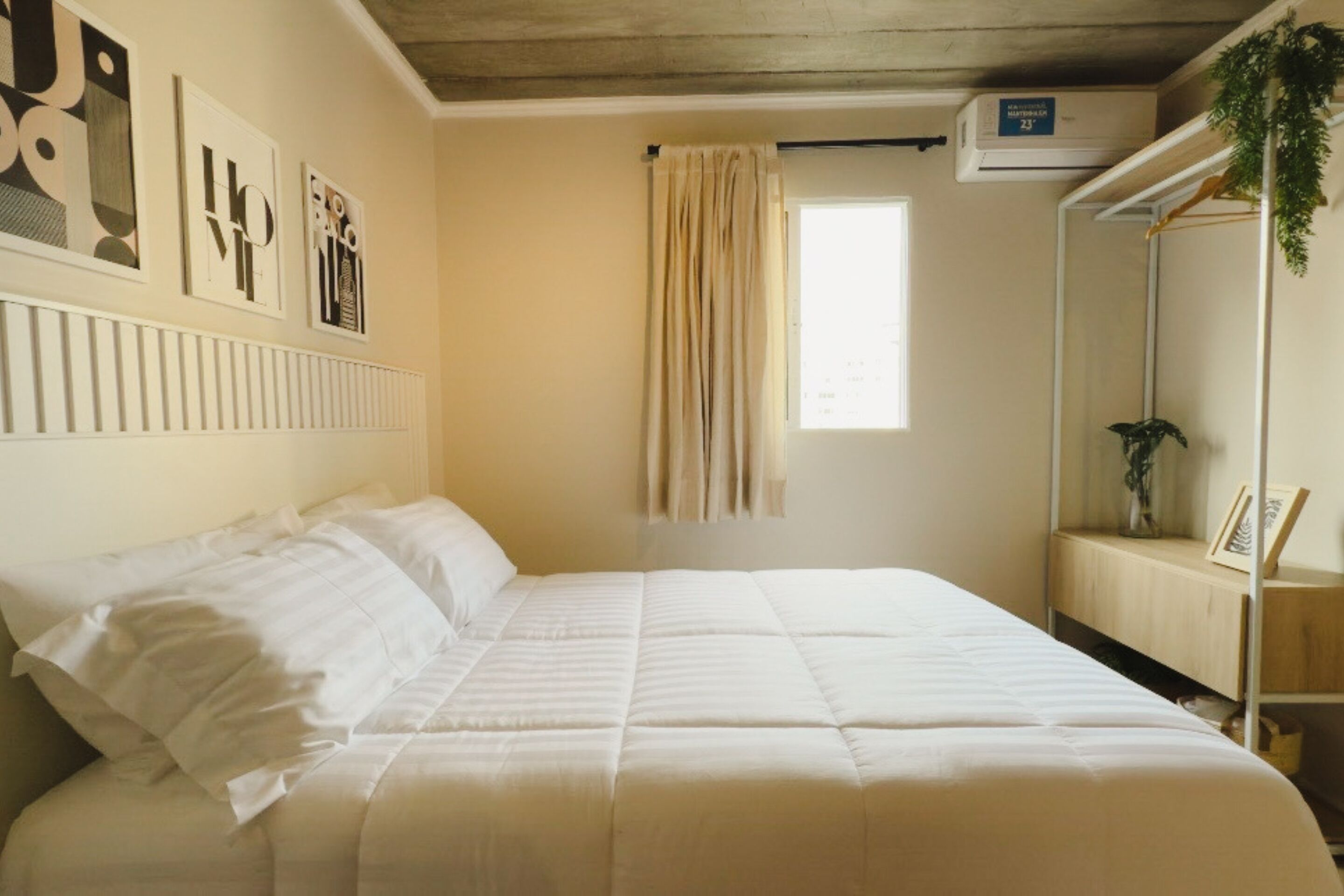A neatly made bed with white bedding, a window with curtains, and a wall-mounted air conditioner.