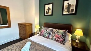 Ein ordentlich gemachtes Bett mit gemusterten Betttextilien, ein Nachttisch aus Holz mit Lampe und zwei gerahmte Bilder an der Wand.