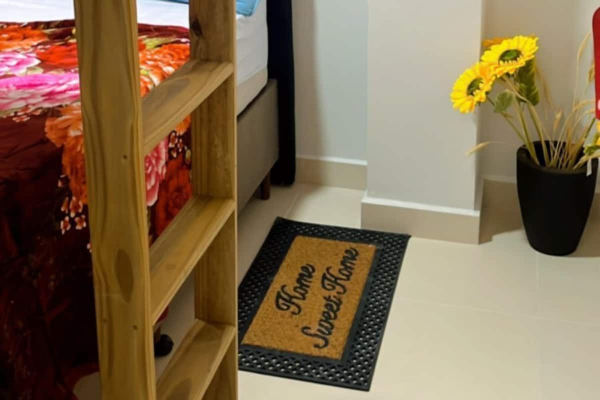 A bunk bed with a red blanket, a blue pillow, and a wooden ladder. There is a black doormat with "Home Sweet Home" written on it. A potted plant with yellow flowers is on the floor.