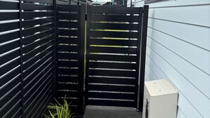 A narrow outdoor corridor with a black gate, a small planter with green plants, and a white utility box against a white building wall. - Brand New Cosy and Convenient Retreat in Taupo  (Taupo)