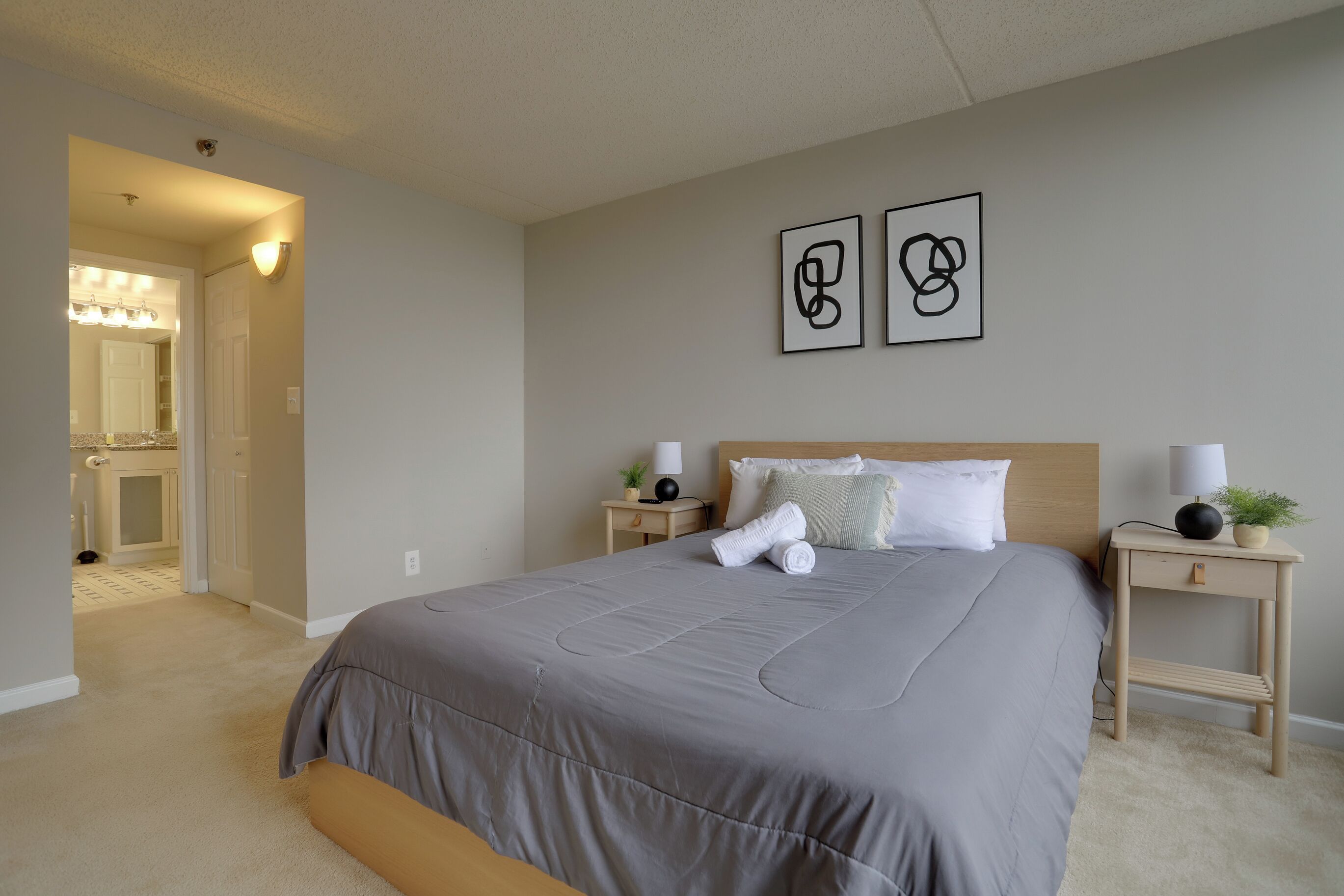 Une chambre avec un grand lit, deux tables de chevet avec des lampes, une petite table de nuit avec une plante, et une porte donnant sur une autre pièce.