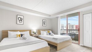 A hotel room with two beds, a nightstand, a window with a city view, and a door. - Crystal City Apartments near Reagan Airport - We Host (Arlington)