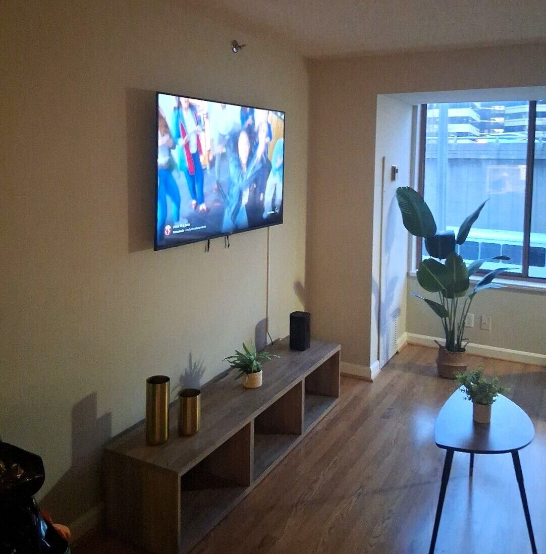 Ett vardagsrum med en platt-TV som är monterad på väggen, en träbänk med dekorativa föremål, ett litet bord och ett stort fönster med utsikt över byggnader.