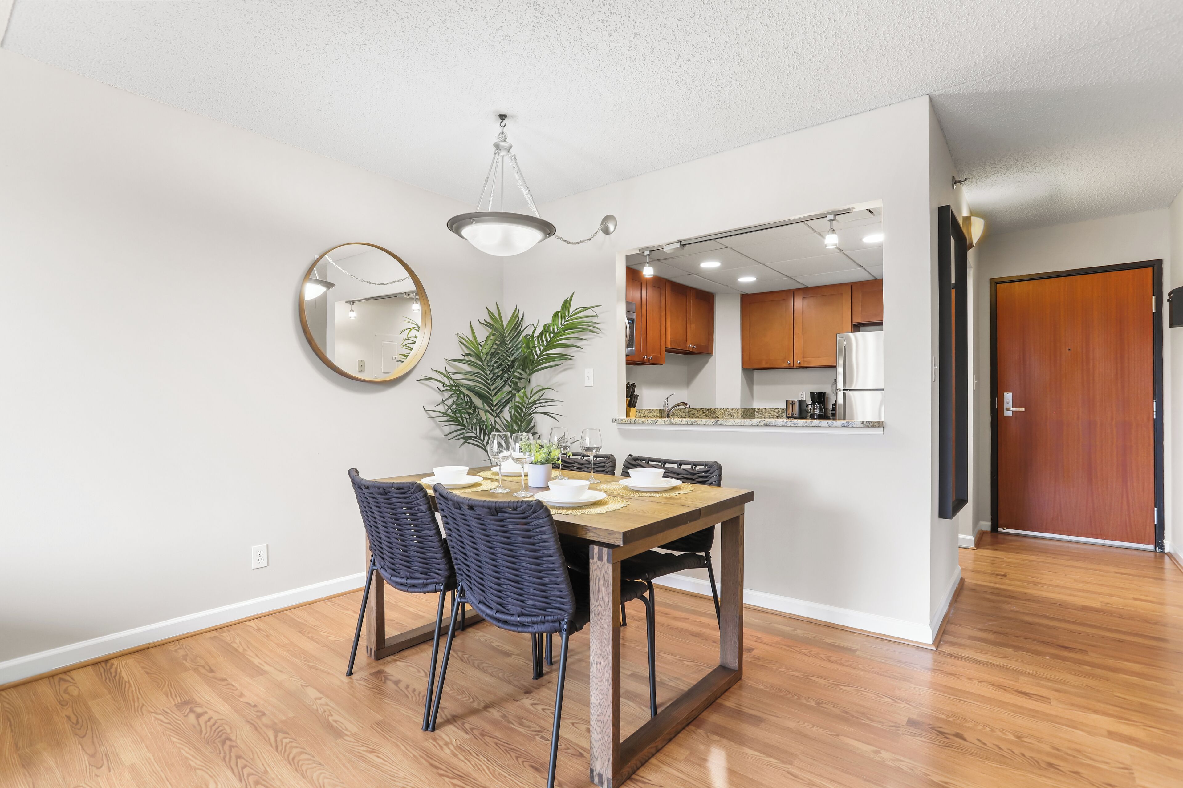 A dining area with a wooden table, chairs, a potted plant, and a kitchen in the background.