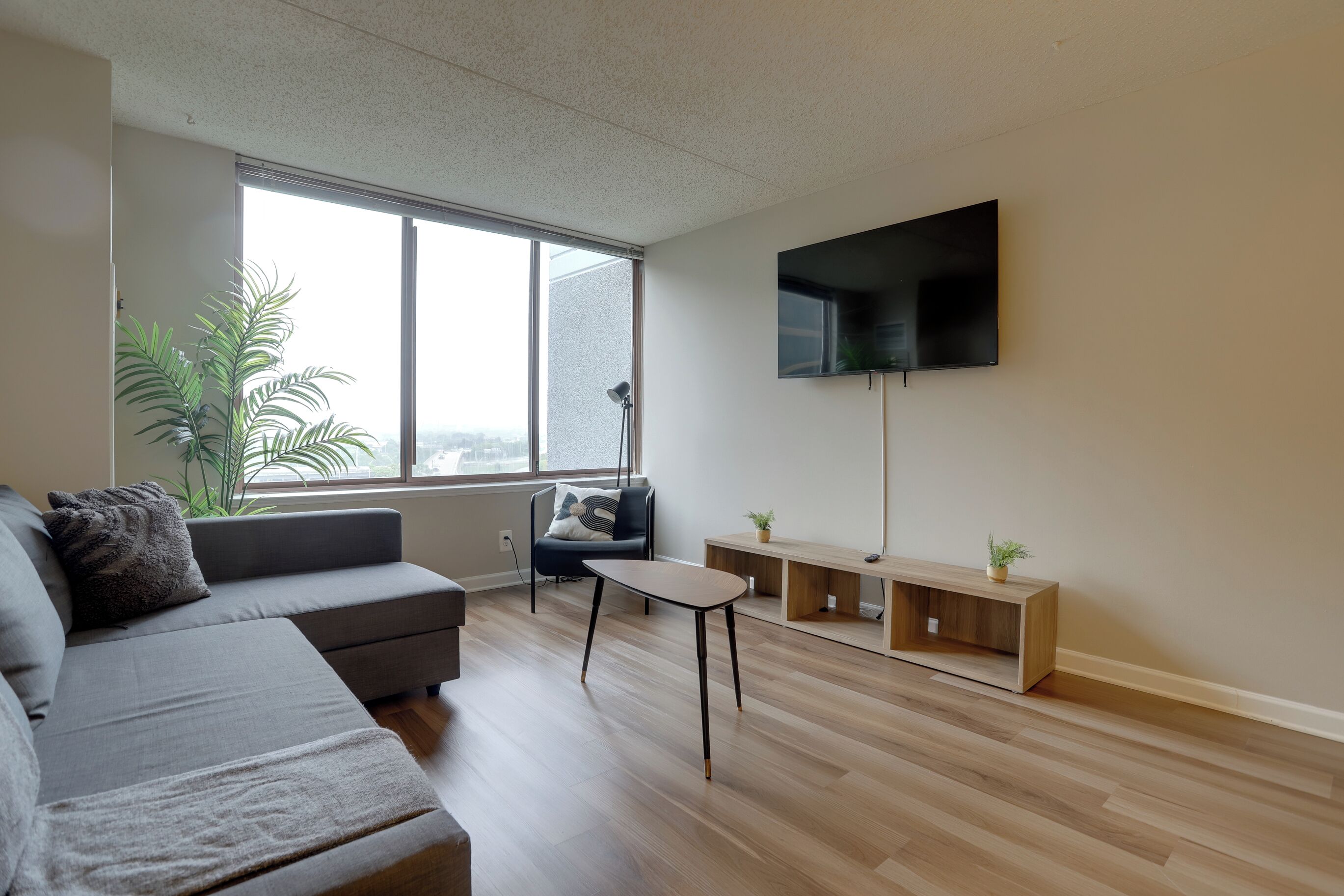 A modern living room with a grey sofa, a small table, a TV mounted on the wall, and a large window with a view.