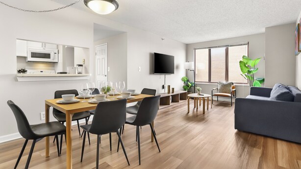 A modern living room with a dining area, wooden flooring, a flat-screen TV, and a large window with plants.
