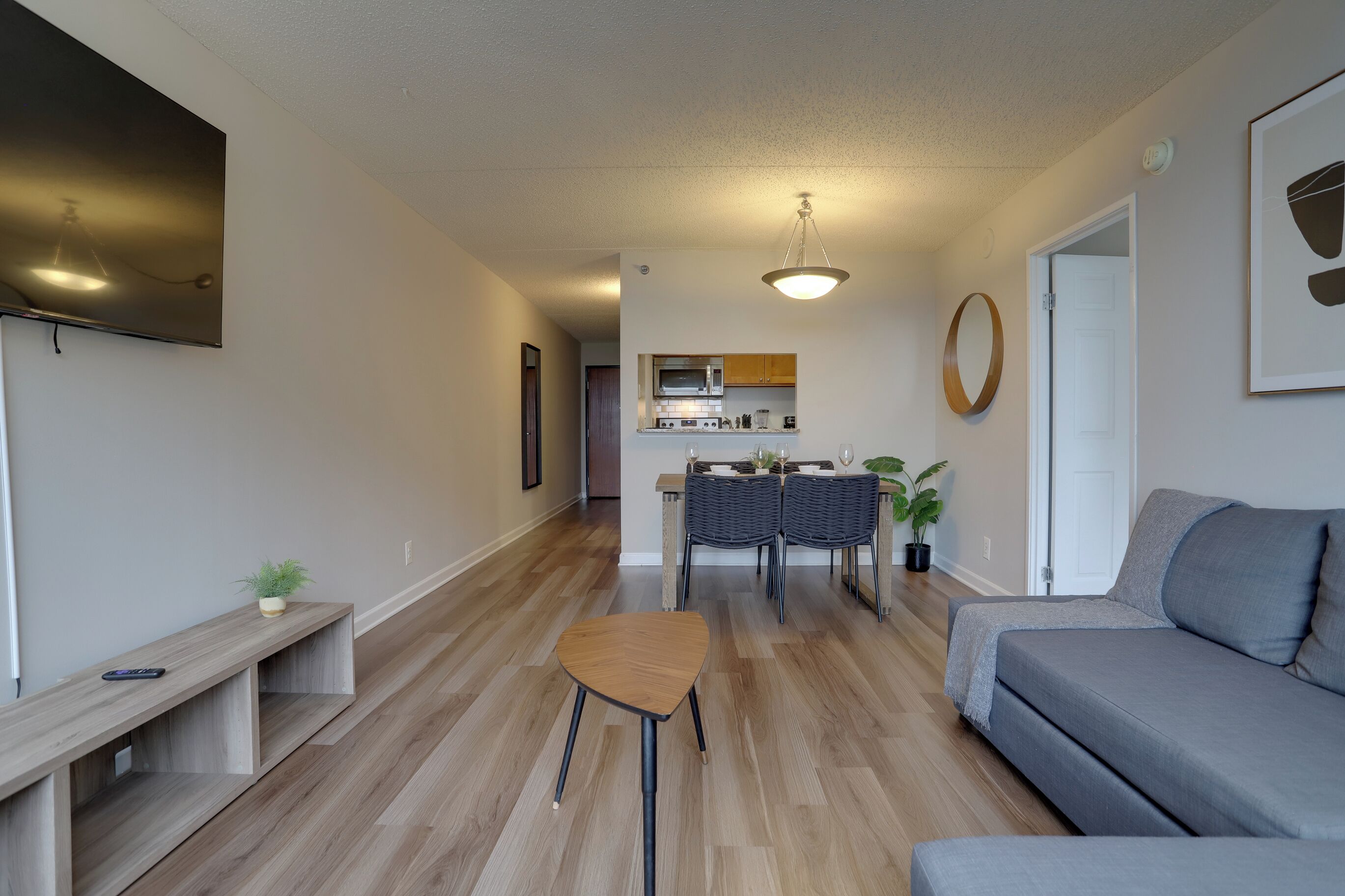 Ein modernes Wohnzimmer mit einem grauen Sofa, einer Holzbank und einem kleinen Hocker. Im Hintergrund ist ein Essbereich mit Tisch und Stühlen zu sehen.