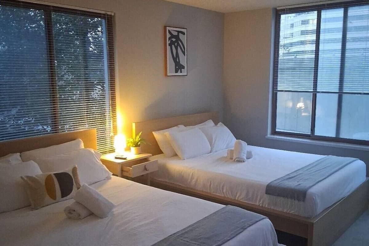 Une chambre d’hôtel avec deux lits, une table de chevet avec une lampe, une fenêtre avec des stores et un tableau encadré au mur.