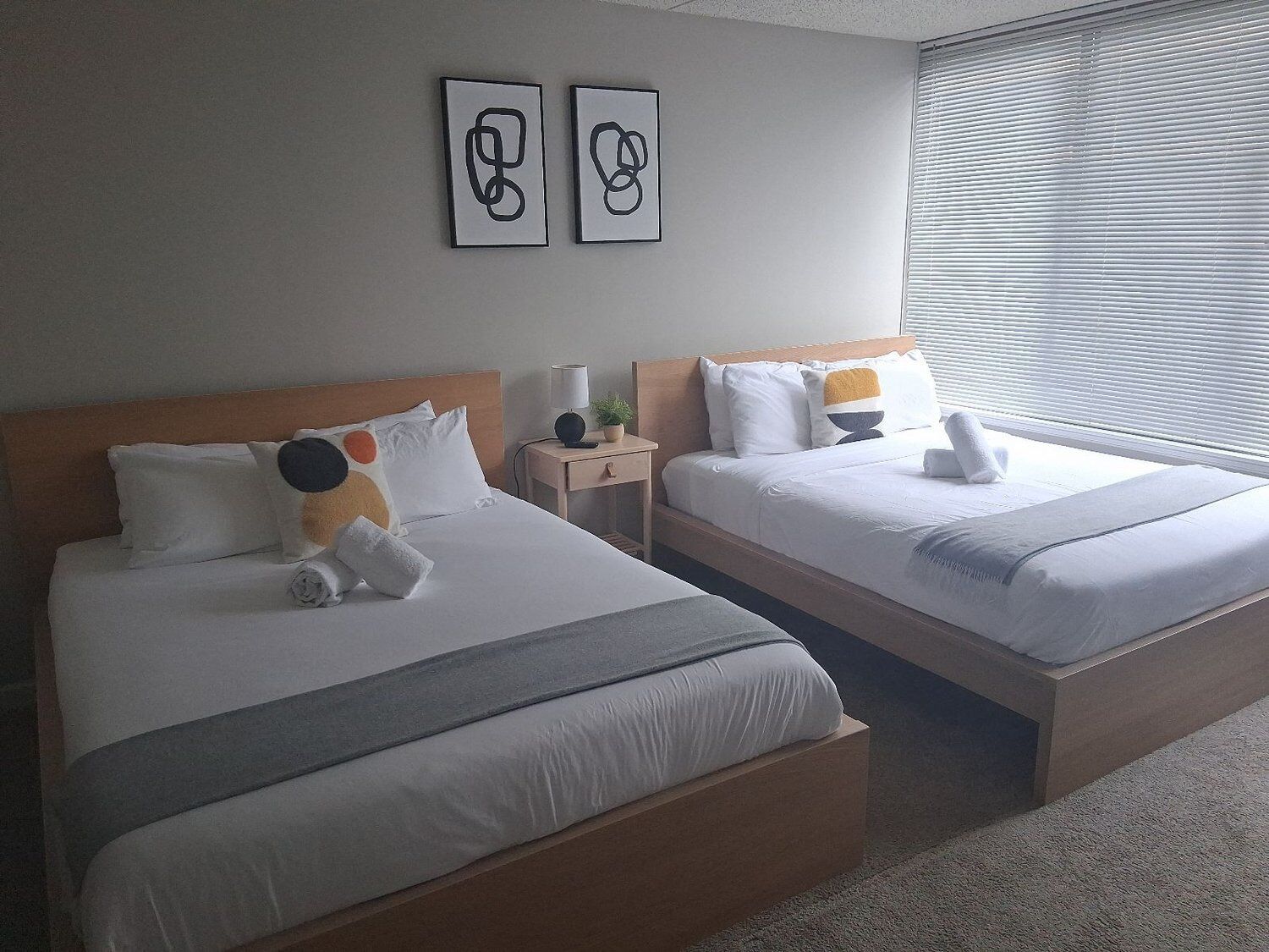 A hotel room with two beds, a nightstand, a lamp, and two wall-mounted artworks.