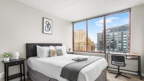 A modern bedroom with a large bed, a desk, and a view of city buildings.