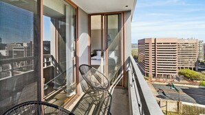 A balcony with two chairs, a bed, and a view of a cityscape and parking lot.