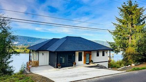 A single-story house with a dark roof, white exterior walls, and a front porch. It is situated by a body of water with a mountain range in the background.