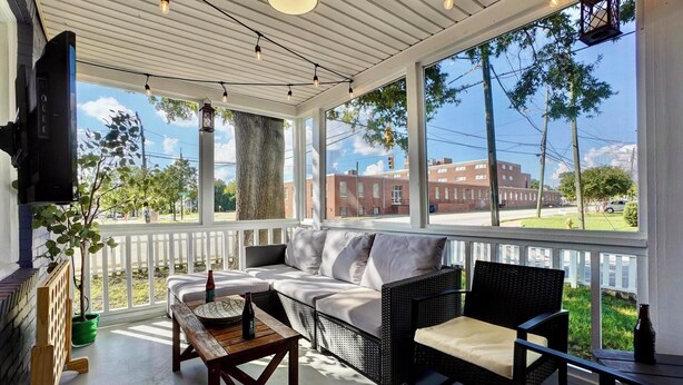 A covered porch with a sofa, chairs, and a table. There is a TV mounted on the wall, a potted plant, and a view of a brick building and street.