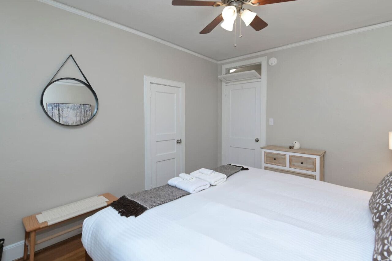 Une chambre à coucher comprenant un lit, un ventilateur de plafond, une table de chevet, une commode, un miroir et une porte.