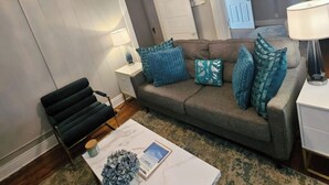 Un salon avec un canapé gris, des coussins bleus, un banc noir, une table basse blanche sur laquelle se trouvent un livre et une plante, et un luminaire suspendu.