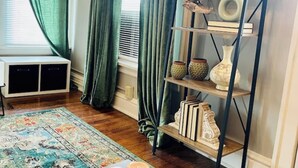 A room with a wooden floor, a patterned rug, a shelf with decorative items, and a window with green curtains. - Espresso Maker & Modern Decor by Arena, University, Hospitals (Huntington)