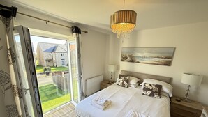 A bedroom with a bed, a painting, a chandelier, and a view of a house through an open door. - Stunning Coastal Cottage - Sleeping 5 in 2 Bedrooms - Pool and Beach Access Included. (Filey)