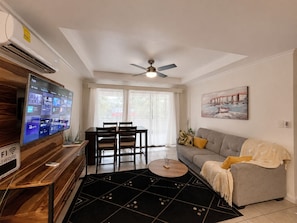 A modern living room with a flat-screen TV, a ceiling fan, a dining area with a table and chairs, and a sofa with a throw blanket.