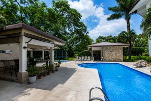 A swimming pool with a covered patio area, lounge chairs, and potted plants.