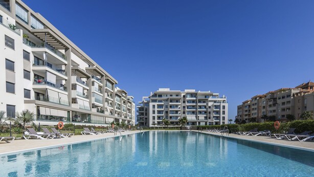 A modern hotel with a central swimming pool, multiple balconies, and a clear blue sky.