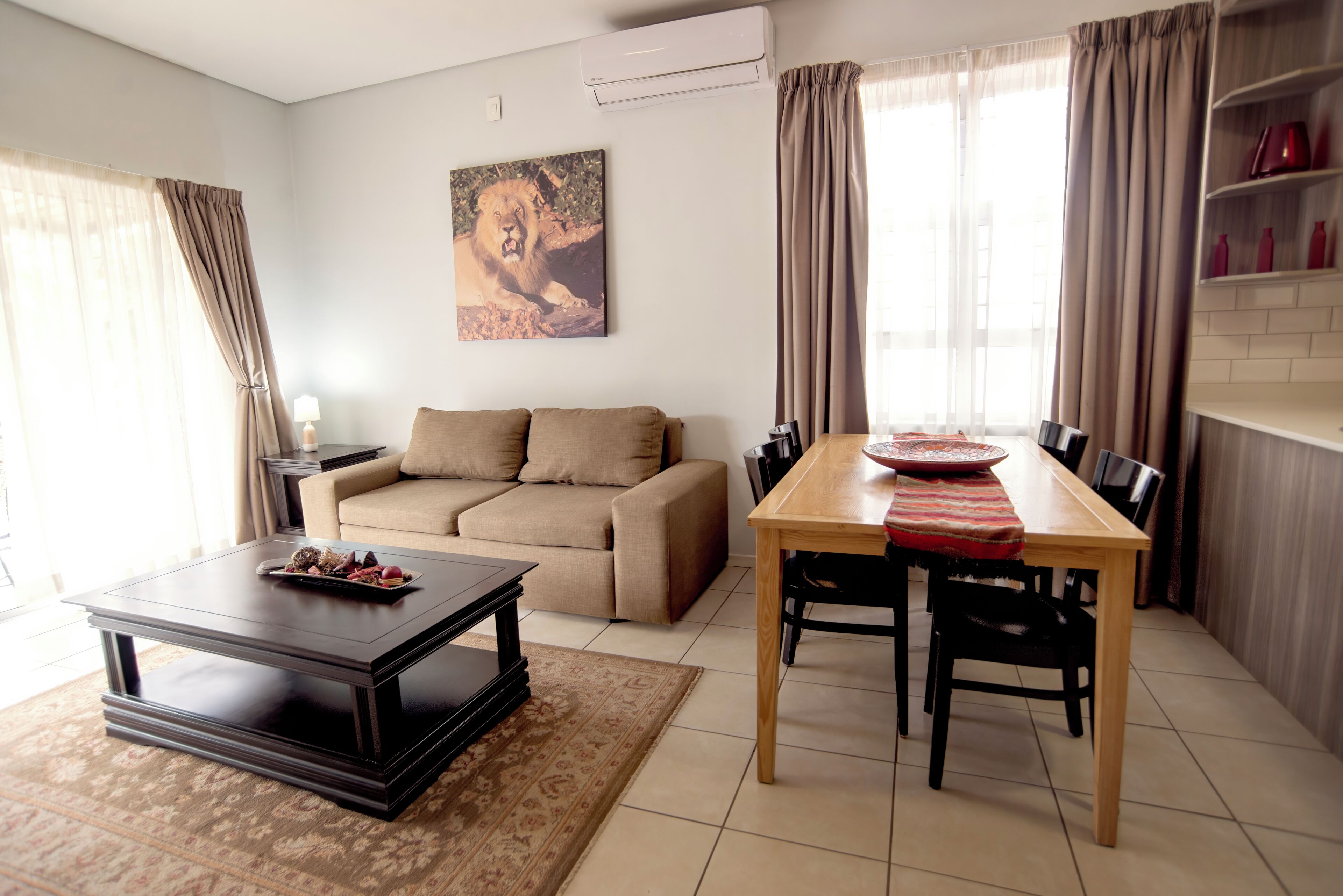 A living room with a sofa, a coffee table, a dining table with chairs, a painting on the wall, and a kitchen area with shelves.