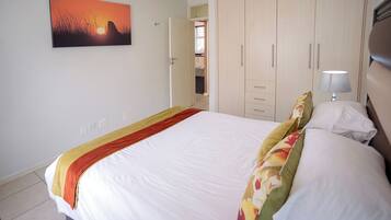 A hotel room with a bed, a wardrobe, a nightstand with a lamp, and a painting of a sunset.