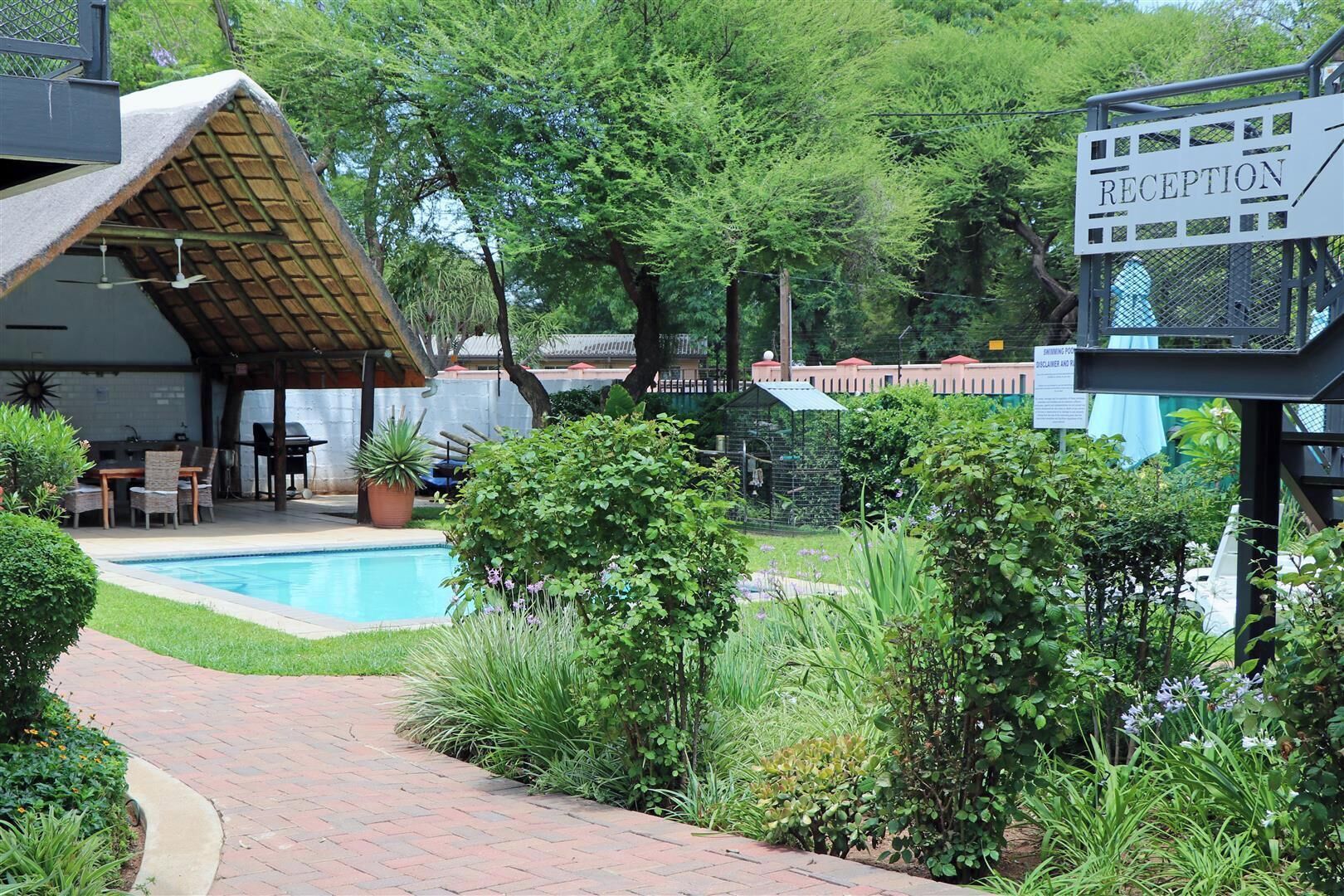 Un área de recepción con un letrero, una piscina, una zona de estar cubierta y abundante vegetación.