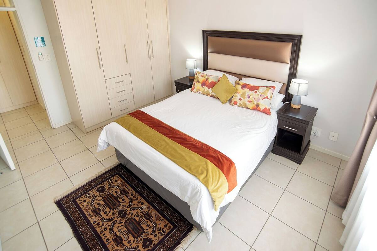A neatly made bed with a patterned rug, a wardrobe, and a bedside table with a lamp.
