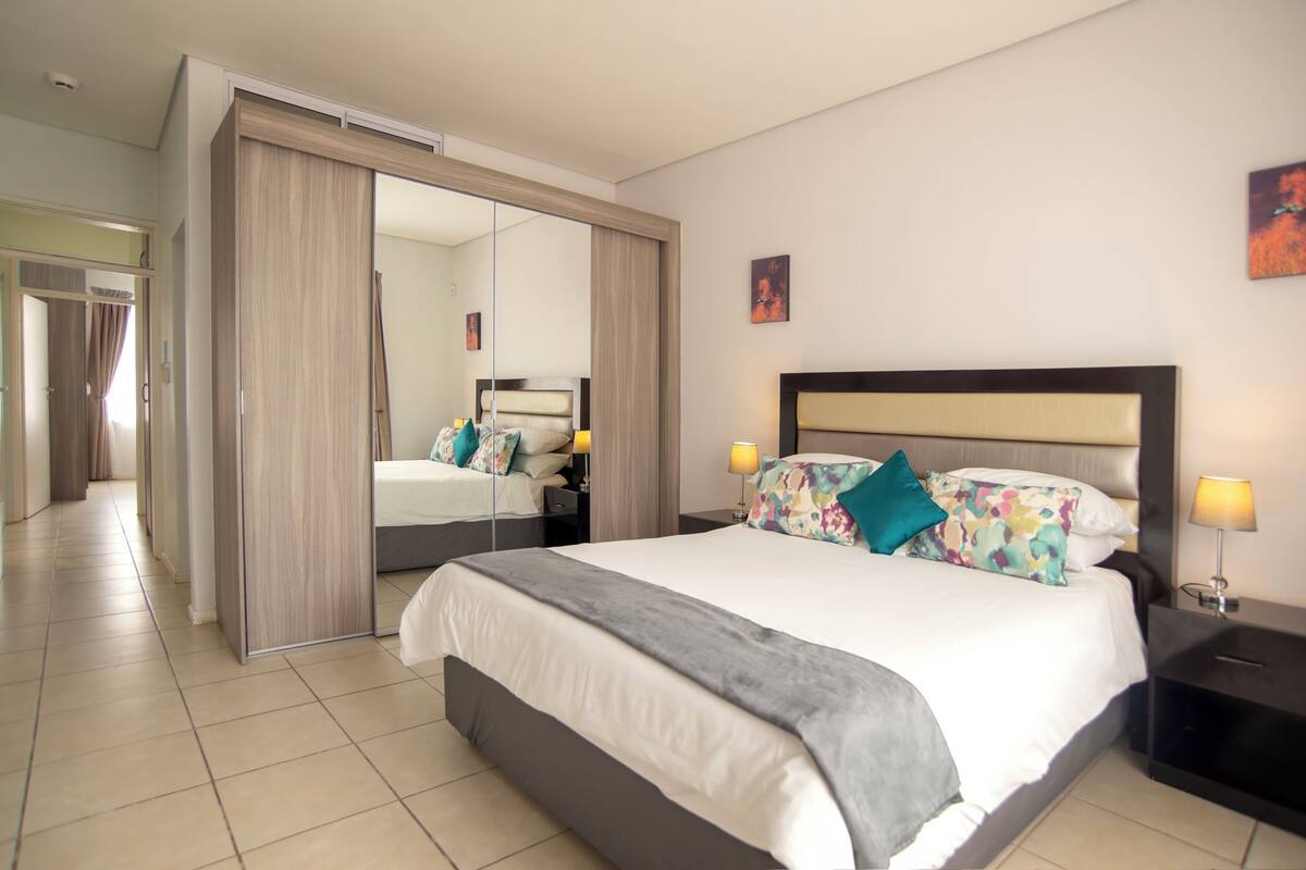 A hotel room with a large bed, a wardrobe, a mirror, and a nightstand with a lamp.