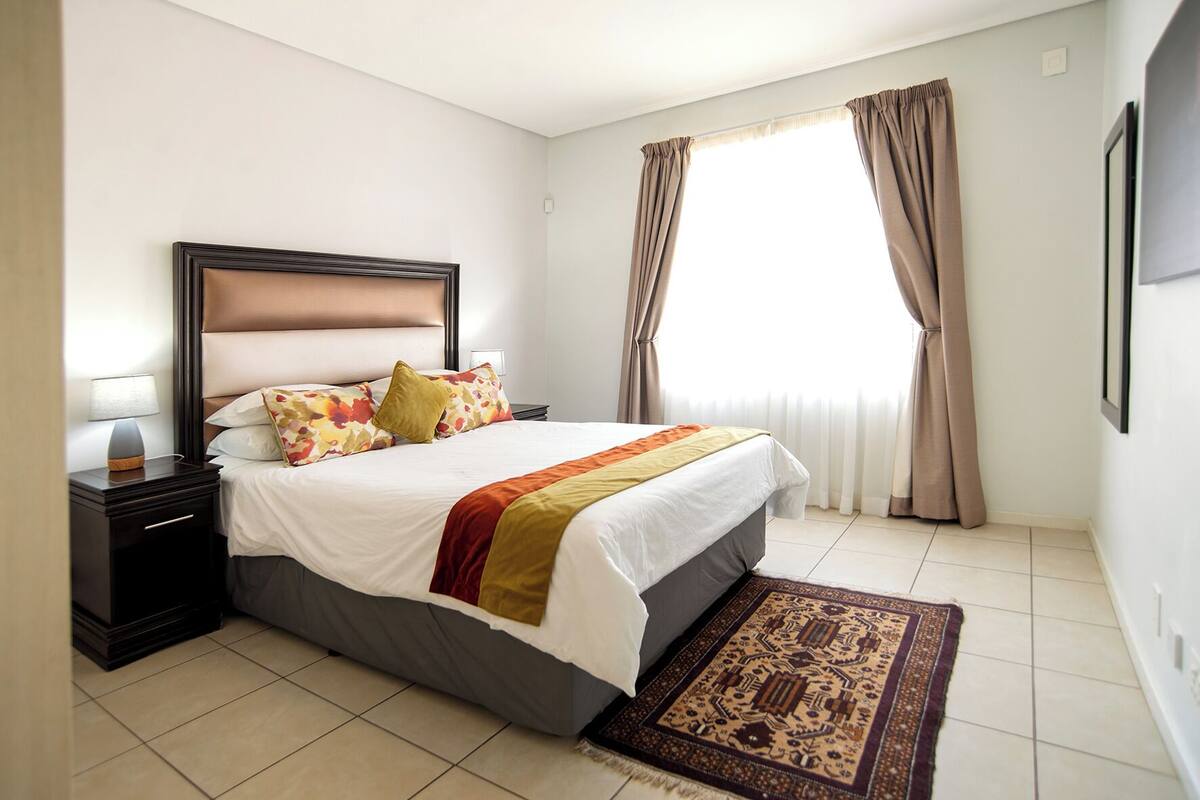 A hotel room with a bed, nightstand, lamp, window with curtains, and a patterned rug.