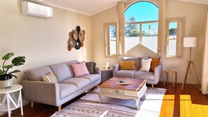 A living room with a grey sofa, a coffee table, a small side table, a floor lamp, and a large window with a view of trees. - Sea Whispers - ocean outlook, walk to 3x beaches (Port Macquarie)