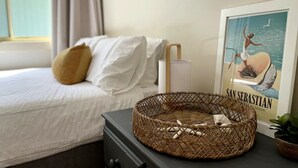A neatly made bed with white linens and a single brown pillow, a wicker basket on a dresser, and a framed picture of a woman on a shell labeled "SAN SEBASTIAN ESPAGNE" on the wall. - Sea Whispers - ocean outlook, walk to 3x beaches (Port Macquarie)