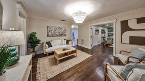 Een ruime woonkamer met een witte bank, een houten salontafel en een grote kroonluchter. Er staat een groene plant en er hangt een ingelijst kunstwerk aan de muur.