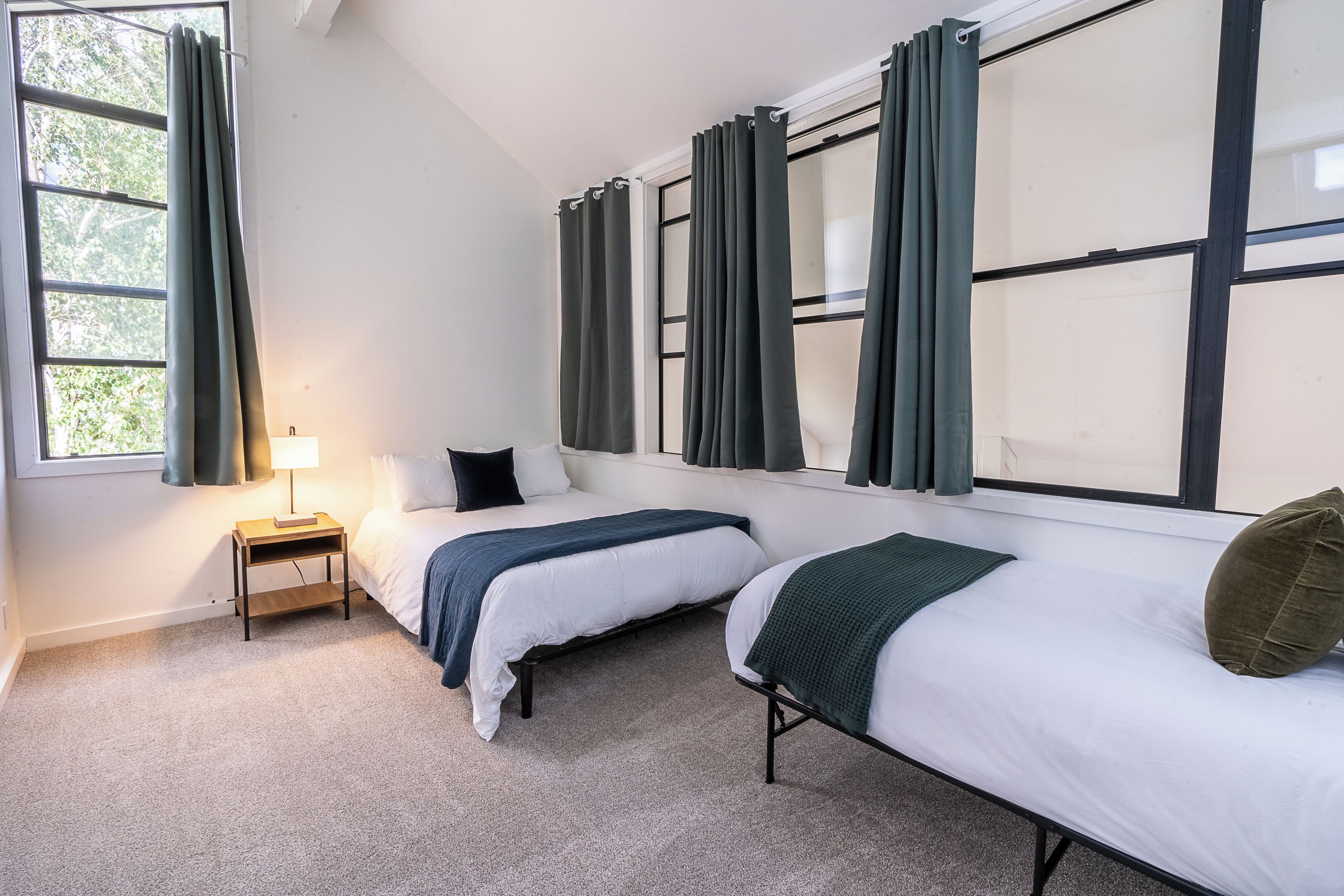 A room with two beds, a nightstand, a lamp, and large windows with curtains.