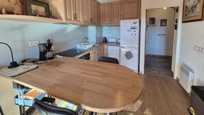 A compact kitchen with wooden cabinets, a built-in dishwasher, a refrigerator, and a small dining area with a wooden table. - Appartement familial au Revard, vue sur le jardin d'enfant et pistes (Les Deserts)