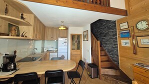 A kitchen with wooden cabinets, a dining table with chairs, a refrigerator, and a staircase with a stone wall. - Appartement familial au Revard, vue sur le jardin d'enfant et pistes (Les Deserts)