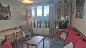 A cozy living room with a sofa, a wooden table, and a chair. There is a balcony with a view of the outdoors.