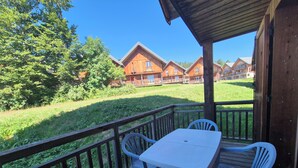 A wooden deck with a table and chairs, overlooking a grassy area and multiple wooden buildings. - Chalet MÖKI , accès piscine sauna *** (Les Deserts)