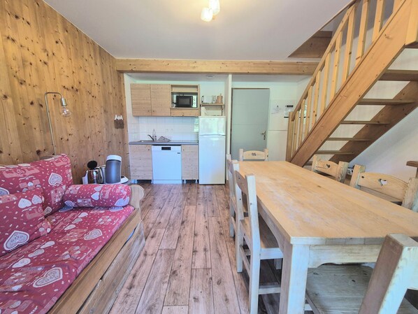A cozy room with a wooden floor, a red patterned sofa, a wooden dining table with chairs, a kitchen area with a microwave, and a staircase. - Chalet MÖKI , accès piscine sauna *** (Les Deserts)