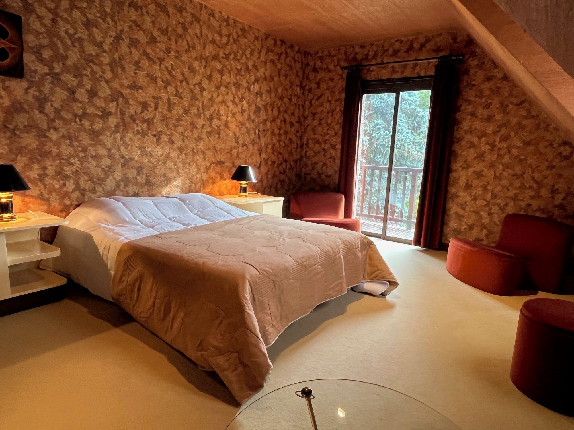 Un dormitorio con una cama grande, dos mesitas de noche con lámparas, una silla roja y un balcón con vistas a árboles.