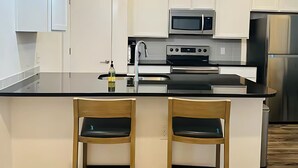 A modern kitchen with white cabinets, stainless steel appliances, and a black countertop. There are two wooden bar stools with black cushions. - Cozy apartment in pleasant Leander with WiFi, AC (Leander)