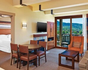 Una camera d'albergo con un letto, una TV, un'area pranzo con sedie, un tavolino e un balcone con vista sul paesaggio.