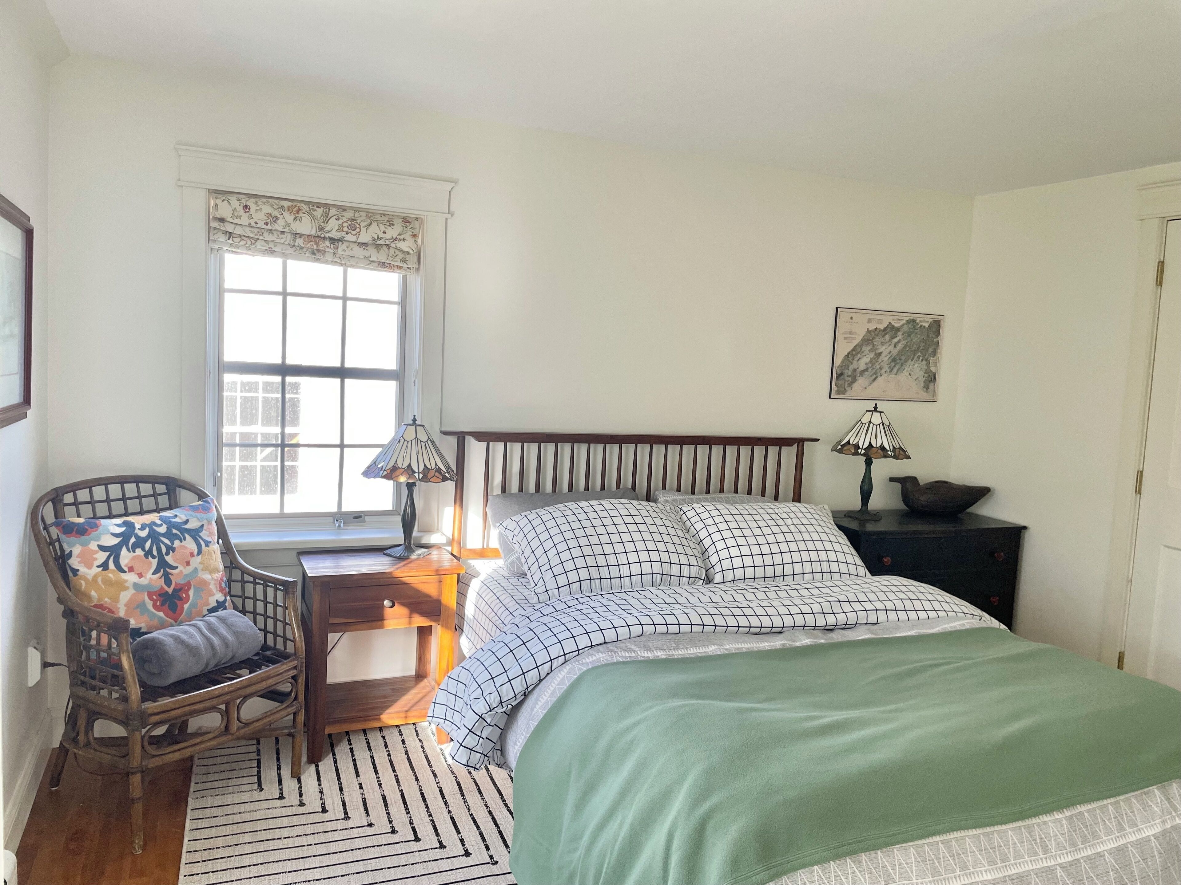A bedroom with a bed, a chair, a nightstand, a lamp, and a window with a floral valance.