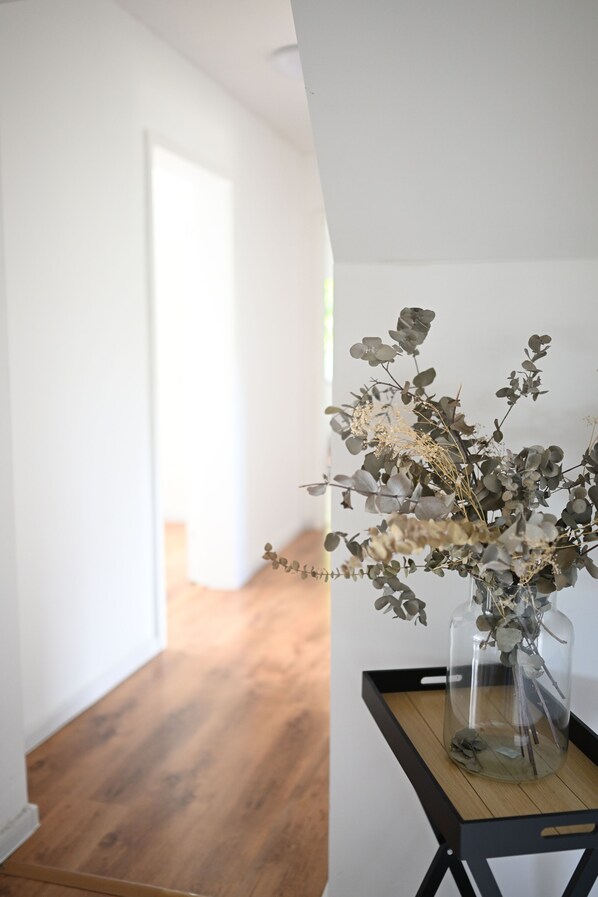 A clear glass vase with dried flowers on a wooden tray against a white wall.
