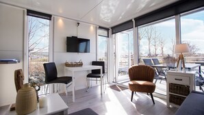 A modern, well-lit room with a large window, a flat-screen TV, a dining table, and comfortable seating.