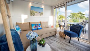 A modern living room with a beige sofa, a blue armchair, and a coffee table with a vase of flowers. A painting of a beach scene hangs on the wall.