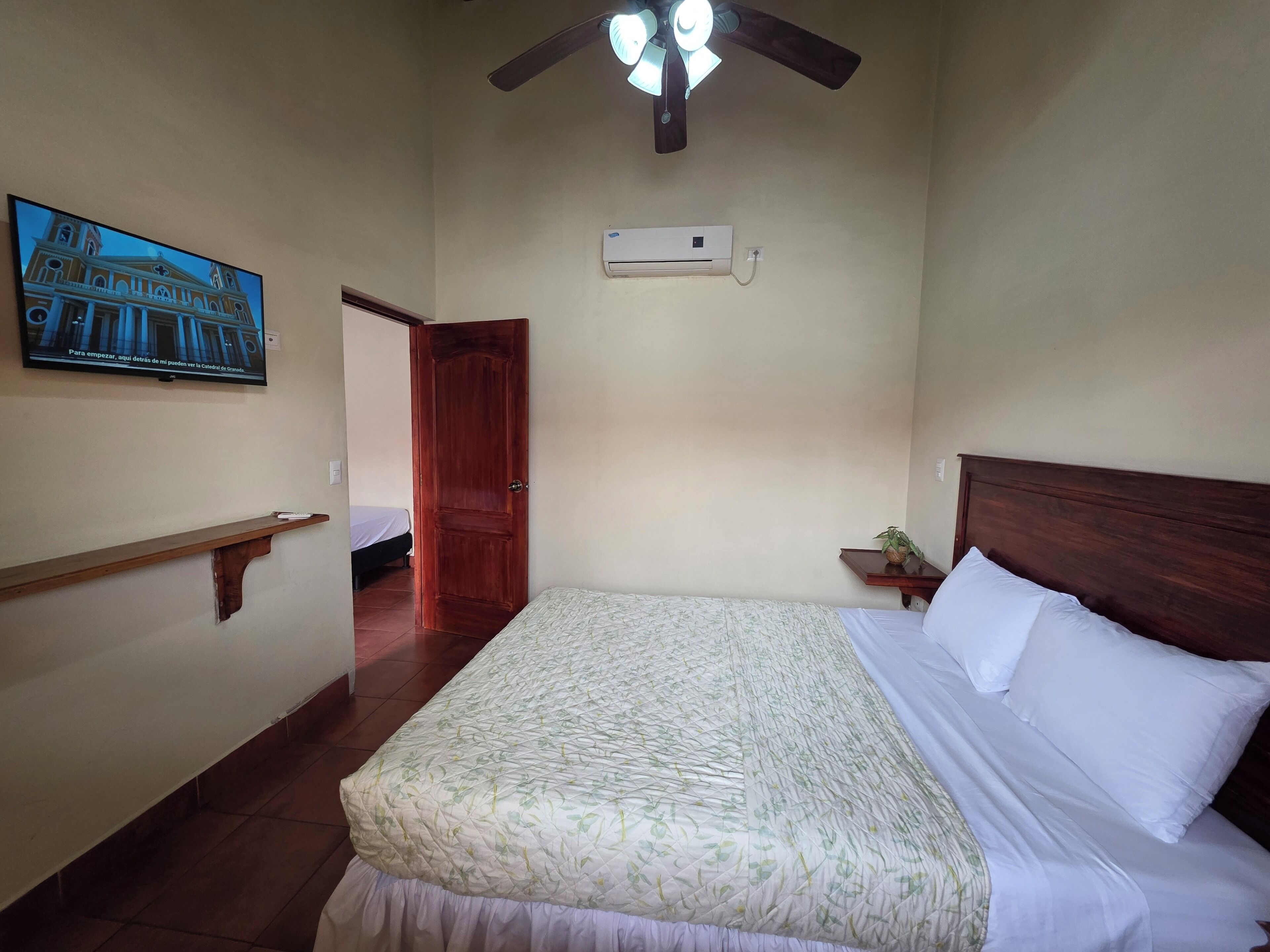 A bedroom with a bed, a ceiling fan, an air conditioning unit, a wooden headboard, a nightstand with a plant, and a wall-mounted TV displaying a building.