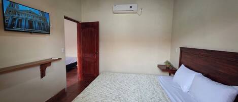 A bedroom with a bed, a ceiling fan, an air conditioning unit, a wooden headboard, a nightstand with a plant, and a wall-mounted TV displaying a building.