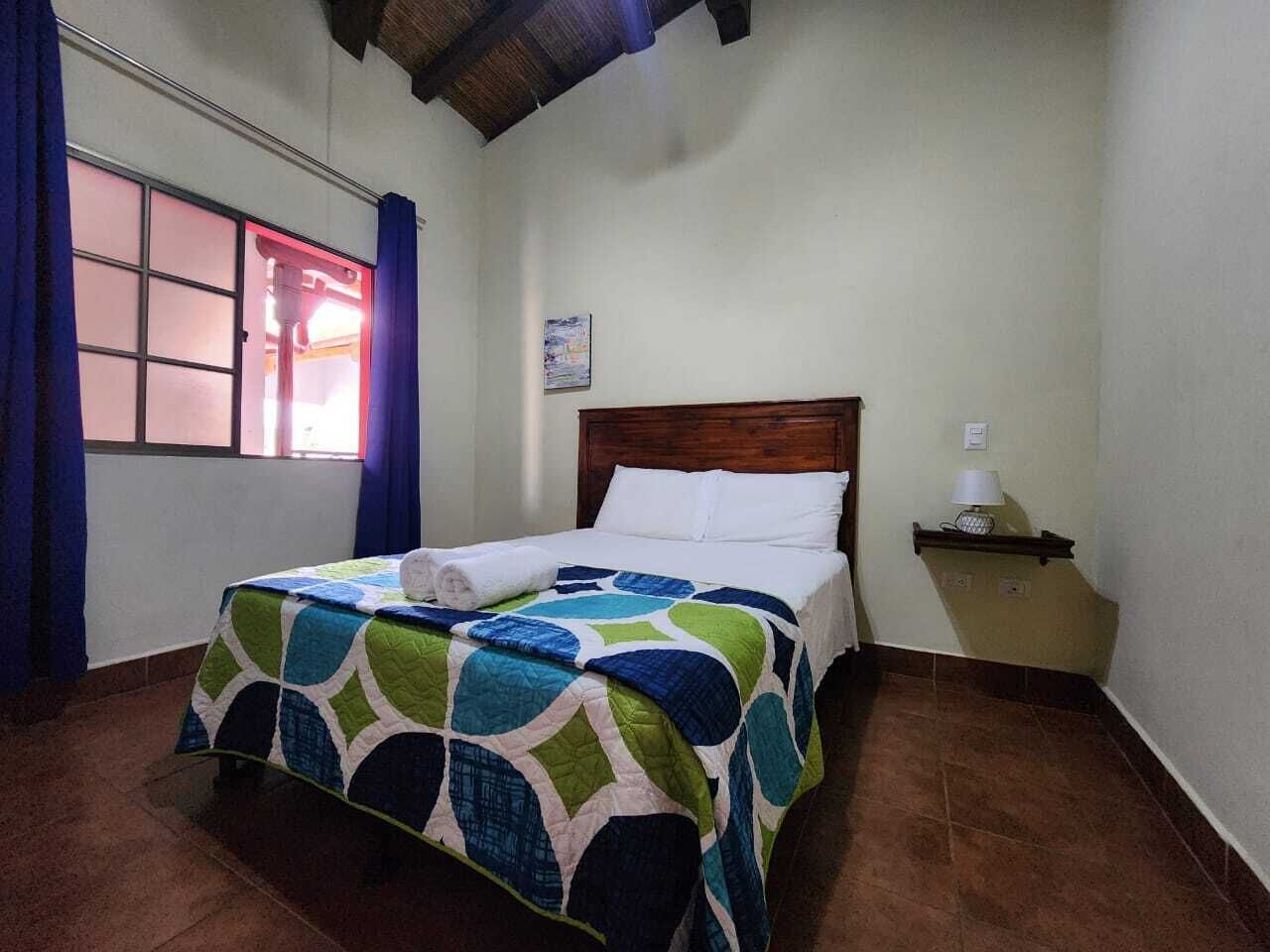 A bedroom with a bed, a nightstand, a window with curtains, and a wall-mounted light.