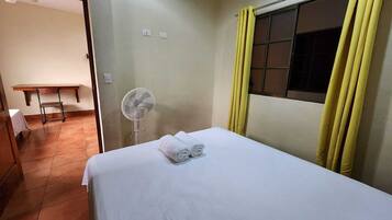 Quarto com cama, ventoinha, janela com cortinas amarelas e mesa com cadeira.